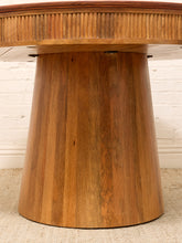 Load image into Gallery viewer, A close-up of a wooden table featuring a fluted round top and a wide, tapered pedestal base made of vertical wood panels, set against a white brick wall and light beige rug.
