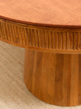 Load image into Gallery viewer, Close-up detail of a round wooden table with a fluted pedestal base and a warm medium-brown wood finish, resting on a light beige textured rug.
