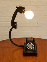 Load image into Gallery viewer, A black rotary telephone turned into a lamp, with its handset detached and shaped like a flexible desk lamp arm, holding a glowing light bulb. The phone sits on a round wooden table against a white painted brick wall.
