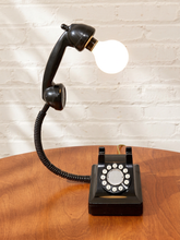 Load image into Gallery viewer, Vintage black rotary telephone converted into a table lamp with raised handset, globe light bulb, coiled cord, and rotary dial
