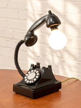Load image into Gallery viewer, A black vintage rotary telephone turned into a table lamp with a glowing light bulb for a shade, displayed on a wooden surface against a white brick wall.
