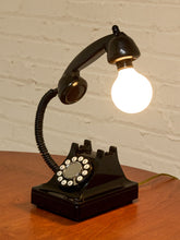 Load image into Gallery viewer, A vintage-style black rotary telephone turned into a table lamp, with the handset detached and shaped like a flexible gooseneck lamp arm holding a glowing light bulb. The lamp sits on a wooden surface against a white painted brick wall.

