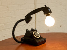 Load image into Gallery viewer, A vintage-style black rotary telephone turned into a table lamp with a glowing light bulb as the shade, set on a wooden table against a white brick wall.
