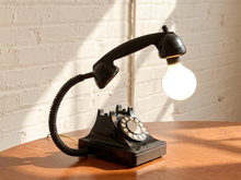 Load image into Gallery viewer, A novelty table lamp shaped like a vintage black rotary telephone, with the handset turned into a light source from a glowing light bulb, set on a wooden table against a white brick wall.
