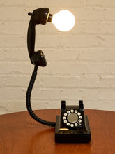 Load image into Gallery viewer, A black rotary telephone turned into a lamp, with the handset detached and connected to a glowing light bulb on a wooden table against a white brick wall.

