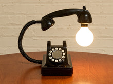 Load image into Gallery viewer, A novelty lamp shaped like a vintage black rotary telephone, with the handset turned into a light source. The lamp sits on a wooden table against a white brick wall, with the bulb glowing brightly.
