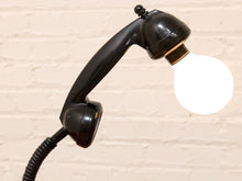 Load image into Gallery viewer, A black vintage-style telephone handset turned into a lamp, with a glowing white bulb where the receiver would be, set against a light beige brick wall.
