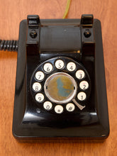 Load image into Gallery viewer, A vintage black rotary telephone with white numbered buttons and a circular dial window, photographed from above on a wooden surface.
