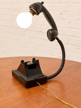 Load image into Gallery viewer, A novelty lamp shaped like a vintage black telephone handset on a base, with a glowing bulb as the light source and a coiled cord, shown on a wooden table against a white brick wall.

