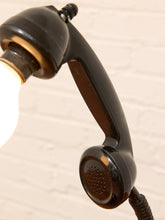Load image into Gallery viewer, A vintage black telephone handset turned into a lamp with a glowing light bulb inserted, set against a light brick wall background.
