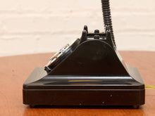 Load image into Gallery viewer, A vintage black rotary telephone with a glossy finish, resting on a wooden table against a white brick wall background.
