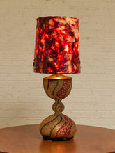 Load image into Gallery viewer, A decorative table lamp with a textured, marbled red and black shade and a sculptural, woven gold and red base, displayed on a wooden table against a white brick wall.
