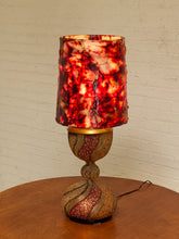 Load image into Gallery viewer, A decorative table lamp with a textured red and orange marbled shade and a matching swirled gold and red base, sitting on a wooden table against a white brick wall.
