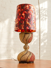 Load image into Gallery viewer, A decorative table lamp with a textured, marbled red and orange shade and a sculptural, woven beige and red base, set on a wooden table against a white brick wall.
