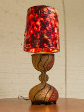 Load image into Gallery viewer, A decorative table lamp with a textured, marbled red and orange glass shade and a matching patterned gold and red base, set on a wooden table against a white brick wall.
