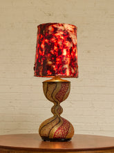 Load image into Gallery viewer, A decorative table lamp with a textured, marbled red and orange shade and a matching patterned gold and red base, sitting on a wooden table against a white brick wall.
