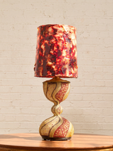Load image into Gallery viewer, A decorative table lamp with a marbled red, orange, and black shade, and a sculptural hourglass-style base in cream, gold, and red mosaic patterns, set on a wooden table against a white brick wall.
