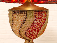 Load image into Gallery viewer, A close-up of a decorative table lamp shade or bowl-like lamp body covered in small glass beads in red, amber, cream, and dark brown, with a warm metallic gold top and base, set against a white brick wall background.
