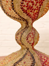Load image into Gallery viewer, Close-up of a sculptural glass vase or decorative sculpture with a bulbous base, a twisted neck, and a flared upper section covered in textured glass beads in cream, red, brown, and gold tones against a light neutral background.
