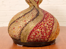 Load image into Gallery viewer, A decorative sculptural vase or lamp base with a bulbous rounded form, covered in textured gold-toned lines and rows of red, amber, and dark brown faceted beads, sitting on a wooden surface against a white brick wall.
