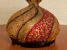 Load image into Gallery viewer, A decorative glass vase with a rounded bulbous body, featuring textured bands of red, amber, and clear glass beads on a dark base, set on a wooden table against a white brick wall.
