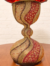 Load image into Gallery viewer, A decorative hourglass-shaped table lamp with a textured, beaded glass body in gold, red, and brown tones, sitting on a wooden surface against a white brick wall.
