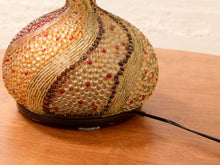 Load image into Gallery viewer, A decorative table lamp with a rounded, bulb-shaped shade covered in textured glass beads and sequins in gold, beige, brown, and red tones, sitting on a wooden table.
