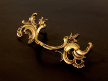 Load image into Gallery viewer, An ornate baroque-style brass drawer pull handle with intricate scrollwork and leaf motifs, displayed on a dark wooden surface.
