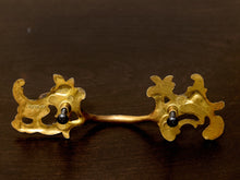 Load image into Gallery viewer, An ornate gold-tone metal hair barrette with decorative scrollwork ends and a central connector, displayed on a dark brown wooden surface.
