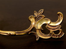 Load image into Gallery viewer, An ornate vintage-style gold metal hardware piece with a decorative leaf and scroll motif, shown close-up on a dark brown wooden surface.
