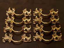 Load image into Gallery viewer, Eight ornate gold-finished metal cabinet pulls arranged in two columns of four on a dark brown wooden surface.

