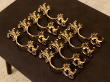 Load image into Gallery viewer, A set of ornate gold-toned metal cabinet pulls with a baroque-style scrollwork design, arranged on a dark brown wooden surface.

