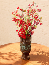 Load image into Gallery viewer, A decorative bronze-colored vase with embossed geometric patterns, filled with red and pink artificial flowers and green stems, set on a round wooden table against a white brick wall.
