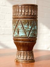 Load image into Gallery viewer, A vintage-style ceramic vase with a textured bronze-brown body, featuring turquoise and white geometric patterns on the center band, set on a wooden surface against a white brick wall.
