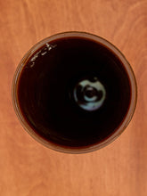 Load image into Gallery viewer, A top-down view of a clear glass containing a very dark brown liquid, set against a warm brown wooden background.
