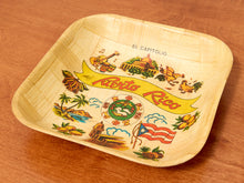 Load image into Gallery viewer, A square, shallow decorative tray made from woven natural fiber, featuring colorful illustrations of Puerto Rican landmarks, flora, and fauna, including the Roto Rico banner and El Capitolio. The tray is photographed on a warm brown wooden surface.
