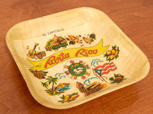 Load image into Gallery viewer, A square, rounded-corner paper plate with a Puerto Rico travel-themed print, displayed on a brown wooden surface. The plate features illustrations of the El Capitulo, Puerto Rican flag, pineapple, and other cultural motifs, with 'EL CAPITULIO' and 'Puerto Rico' printed on it.
