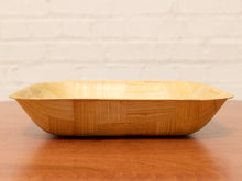 Load image into Gallery viewer, A rectangular wooden serving bowl with a woven bamboo pattern on the exterior and a smooth light wood interior, resting on a wooden table against a white brick wall background.

