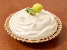 Load image into Gallery viewer, A ceramic pie dish with a cream-colored whipped-look top and a fluted beige rim, topped with a small yellow lemon and green leaf decoration, set on a wooden surface.
