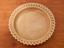 Load image into Gallery viewer, A round ceramic plate with a scalloped rim, shown from a top-down view on a warm brown wooden surface.
