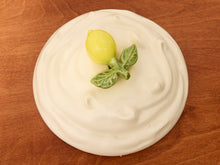 Load image into Gallery viewer, A round white plate or dish with a swirled, whipped-like surface, topped with a small yellow lemon and green leaf decoration, set on a brown wooden surface.
