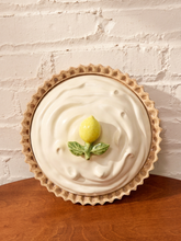 Load image into Gallery viewer, A ceramic pie dish shaped like a lemon tart, featuring a beige fluted rim, a cream-colored swirl pattern, and a small yellow lemon and green leaf detail on top, displayed on a wooden surface against a white brick wall.
