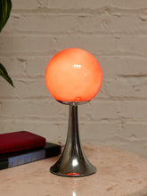 Load image into Gallery viewer, A modern table lamp with a glowing orange spherical shade on a polished silver metal base, set on a stone tabletop next to stacked books against a white brick wall.
