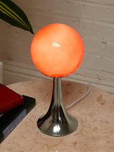 Load image into Gallery viewer, A modern table lamp with a glowing orange spherical shade on a polished silver metal flared base, set on a light stone tabletop with a white brick wall background.
