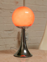 Load image into Gallery viewer, A modern table lamp with a glowing orange spherical shade on a polished chrome stand, set on a light-colored surface against a brick wall background.
