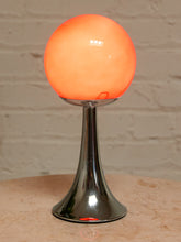 Load image into Gallery viewer, A modern table lamp with a glowing orange spherical shade on a polished chrome flared base, set on a light stone surface against a white brick wall.
