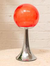 Load image into Gallery viewer, A modern table lamp featuring a translucent orange-red glass globe on a polished chrome flared base, set against a light brick wall background.
