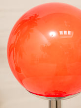 Load image into Gallery viewer, A close-up of a glossy translucent orange-red spherical lamp globe mounted on a chrome stand, with reflections of the room interior visible on its smooth surface.
