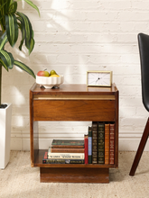 Load image into Gallery viewer, A mid-century modern wooden nightstand with a single drawer and open shelving, styled with books, a small clock, and fruit, set against a white brick wall.
