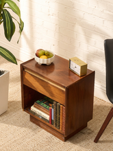 Load image into Gallery viewer, A mid-century modern wooden nightstand with a single drawer and open shelf, styled with a bowl of fruit, a small clock, and books in a bright room with a white brick wall.
